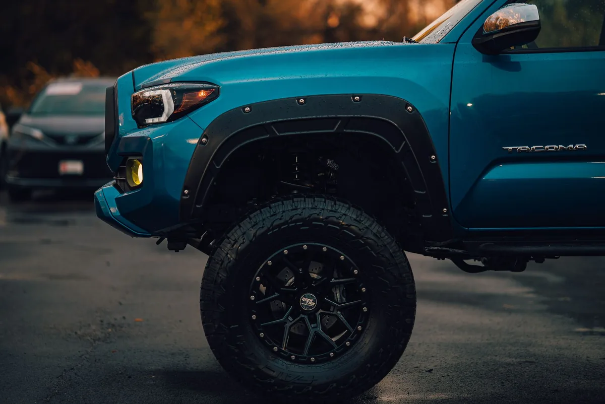 Black aftermarket wheel and all-terrain tire on a lifted blue Tacoma at dusk